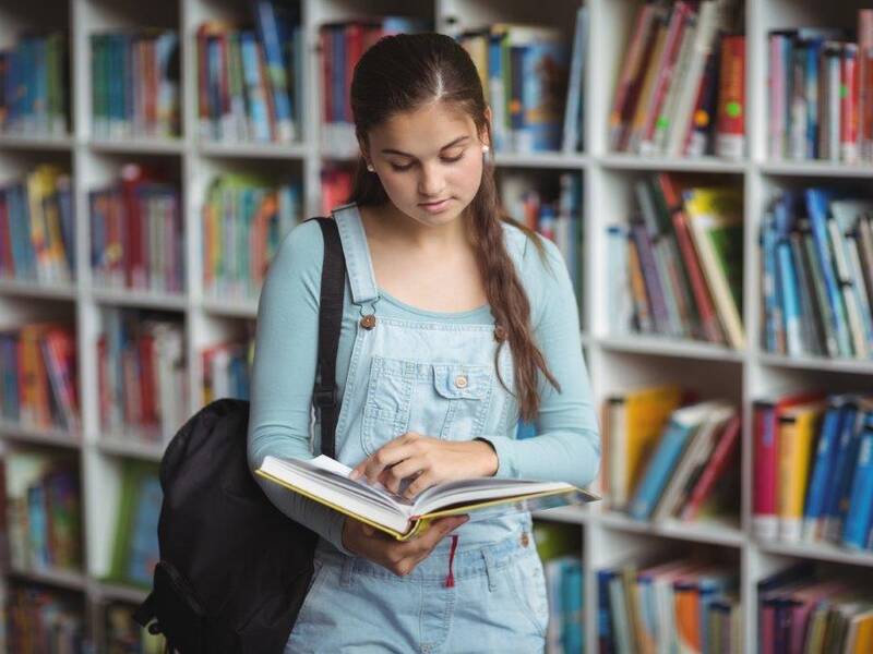 Ein Mädchen liest in einem aufgeschlagenen Buch das sie in der Hand hält und steht vor einem Bücherregal