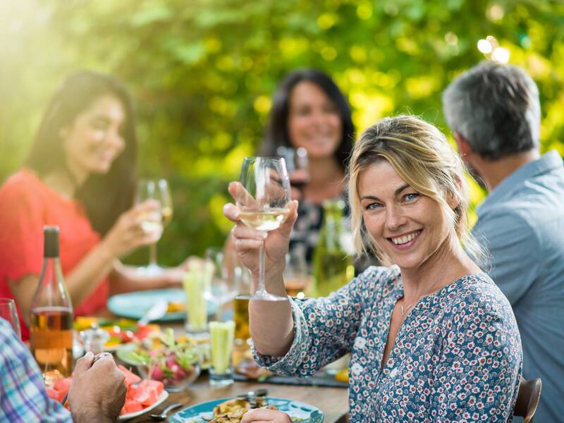 Eine Gruppe von Menschen sitzt draußen an einem Tisch, genießt Essen und Getränke, wobei eine lächelnde Frau im Vordergrund ein Glas Wein hebt.