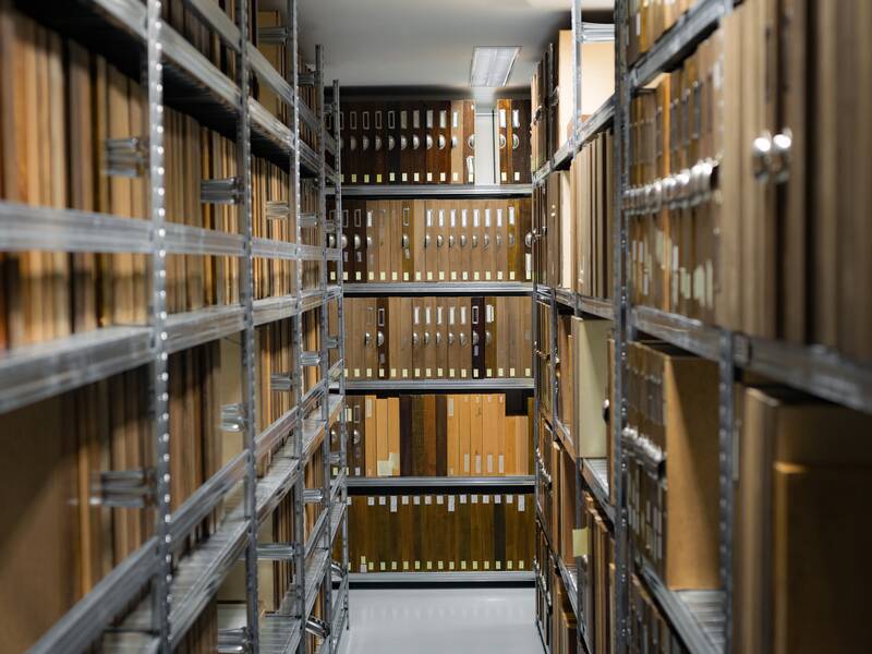 Ein langer, schmaler Gang in einem Archiv mit hohen Regalen, die dicht mit Aktenordnern und Papierdokumenten befüllt sind.