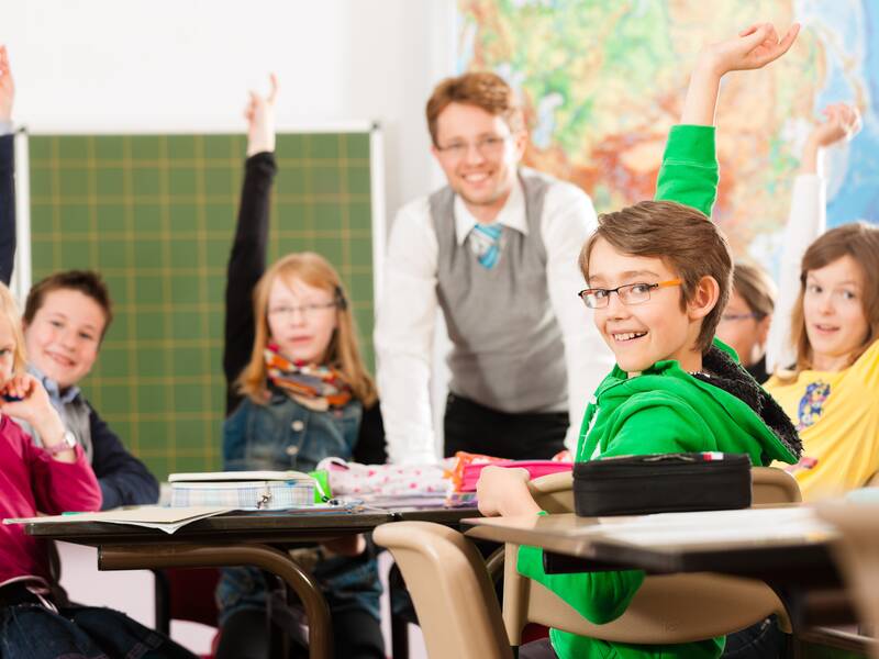 Ein Lehrer und mehrere Schüler in einem Klassenzimmer, von denen einige fröhlich die Hand heben.