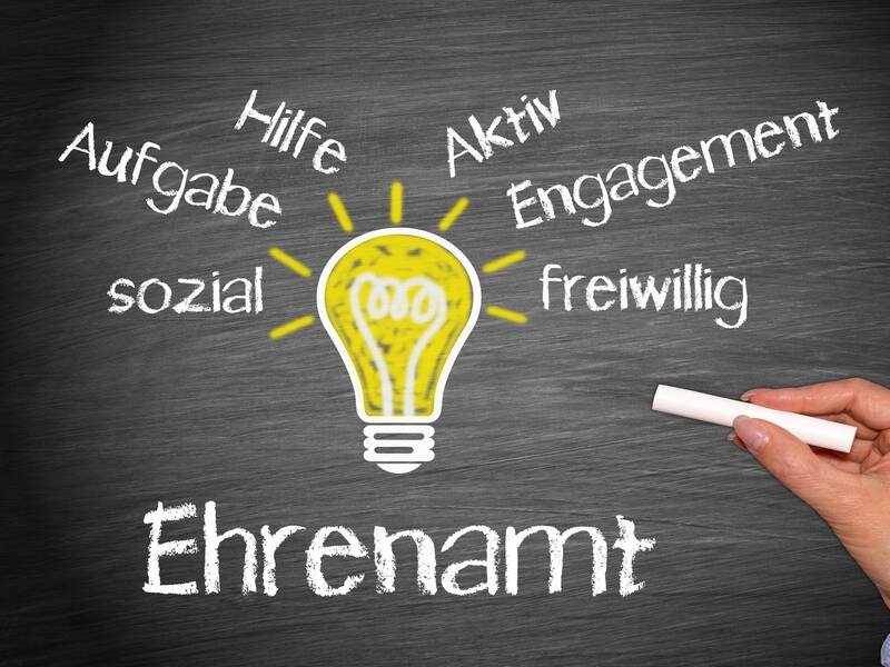 Auf einer Tafel steht das Wort "Ehrenamt" in großen Buchstaben, umgeben von Begriffen wie "Hilfe", "Aufgabe", "sozial", "freiwillig", "Engagement" und "aktiv", die eine gelb gezeichnete Glühbirne umkreisen.