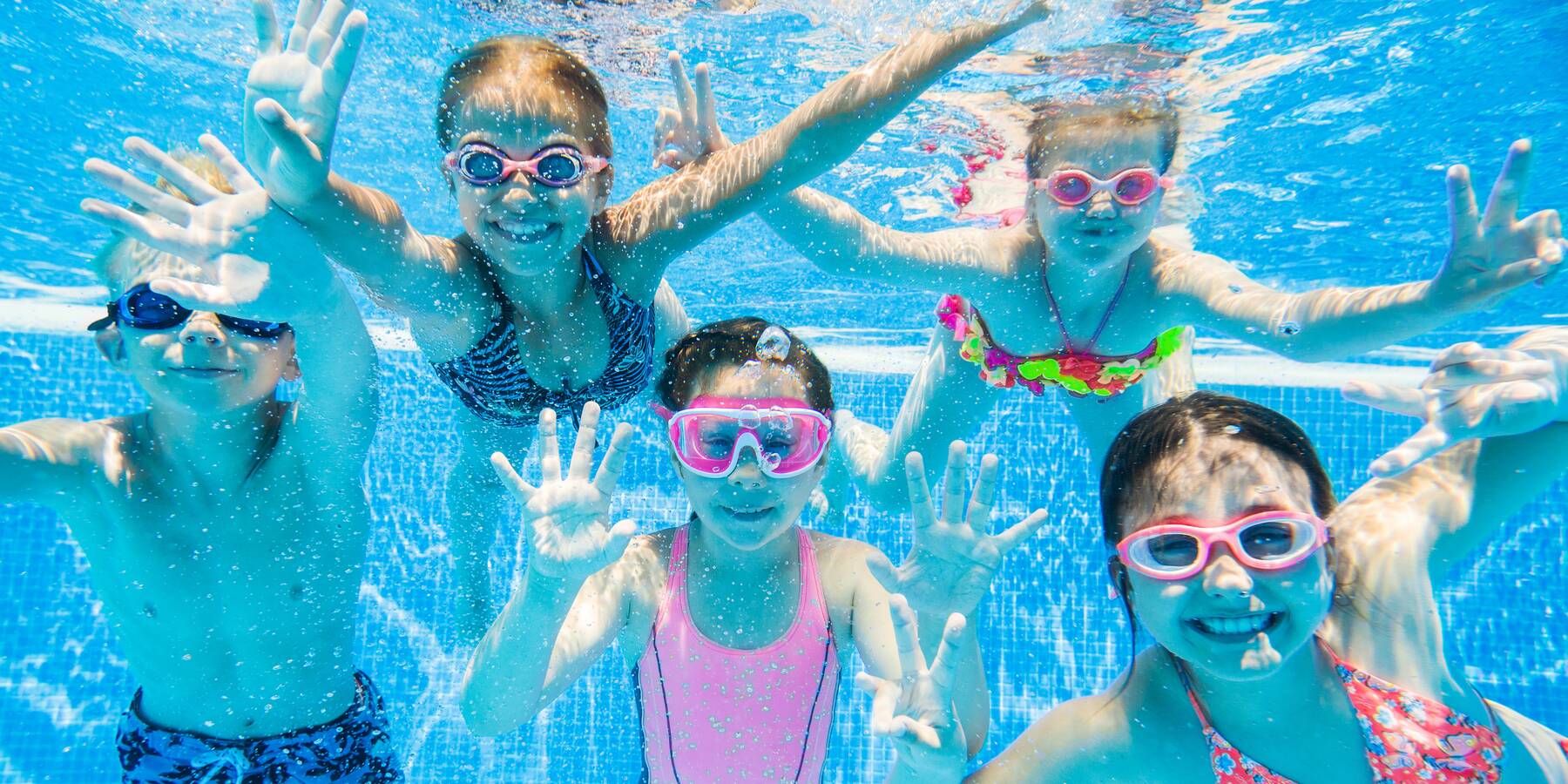 Fünf Kinder mit Taucherbrillen schwimmen unter Wasser in einem Pool und winken fröhlich in die Kamera.