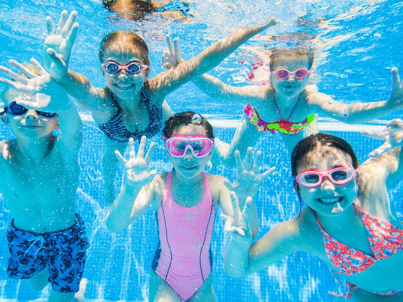Fünf Kinder mit Taucherbrillen schwimmen unter Wasser in einem Pool und winken fröhlich in die Kamera.