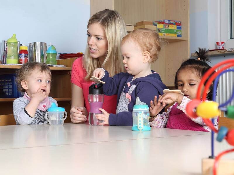 Eine Erzieherin sitzt mit drei kleinen Kindern an einem Tisch, während eines der Kinder einen Trinkbecher hält.