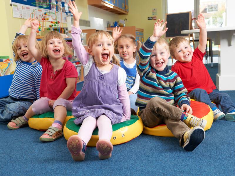 Eine Gruppe von sechs fröhlichen Kindern sitzt nebeneinander im Kreis auf bunten Kissen und hebt lachend die Hand.