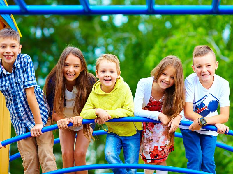 Fünf fröhliche Kinder lehnen sich auf einem Spielplatz an eine blaue Kletterstruktur.
