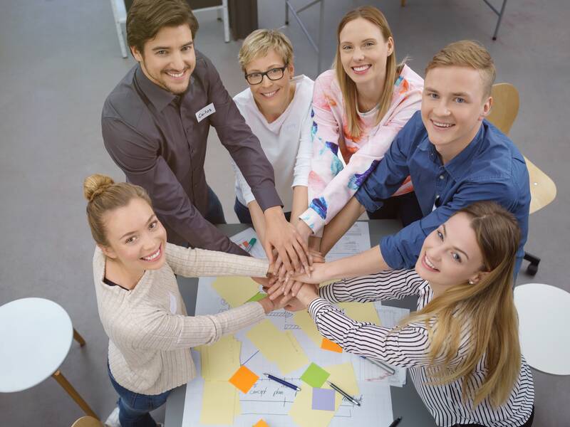 Eine Gruppe von sechs lächelnden Menschen steht um einen Tisch und legt ihre Hände übereinander, als Zeichen der Zusammenarbeit.