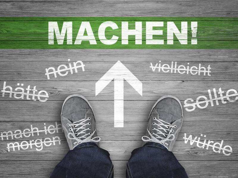 Ein Paar Schuhe steht auf einem Holzboden, umgeben von durchgestrichenen Wörtern wie "hätte" und "vielleicht", während ein großer Pfeil und das Wort "MACHEN!" in grüner Farbe darüber angezeigt sind.