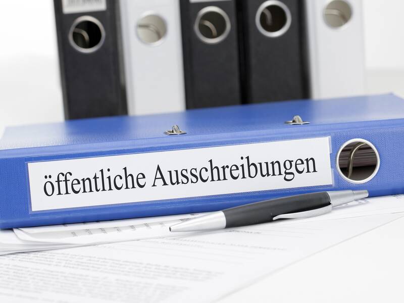 Ein blauer Ordner mit der Aufschrift "öffentliche Ausschreibungen" liegt auf einem Stapel von Dokumenten, daneben ein Kugelschreiber, mit weiteren Ordnern im Hintergrund.