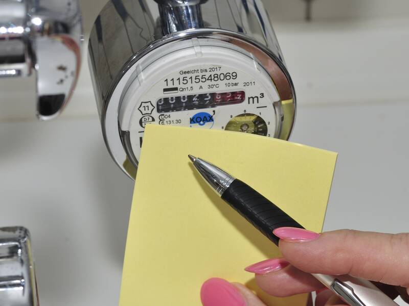 Eine Hand mit rosa lackierten Nägeln hält einen Stift und einen gelben Notizblock neben einen Wasserzähler, auf dem Zahlen abgelesen werden.