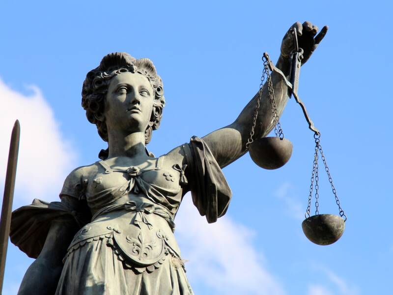 Eine Statue der Justitia mit verbundenen Augen hält eine Waage in der rechten Hand und ein Schwert in der linken, vor einem blauen Himmel.