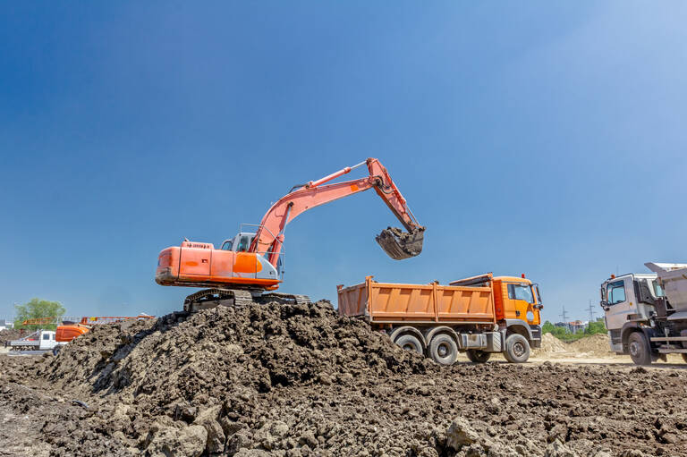 Ein orangefarbener Bagger lädt Erde in einen orangefarbenen Lastwagen auf einer Baustelle unter klarem, blauem Himmel.