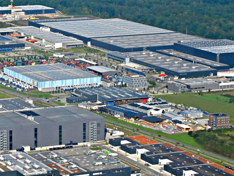 Luftaufnahme eines großen Industrie- und Gewerbegebiets mit mehreren großen Hallen und Gebäuden, umgeben von Feldern und angrenzend an eine Stadtlandschaft im Hintergrund.