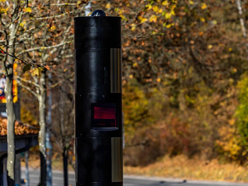 Eine schwarze Blitzersäule. Im Hintergrund sind herbstliche Bäume und Sträucher.