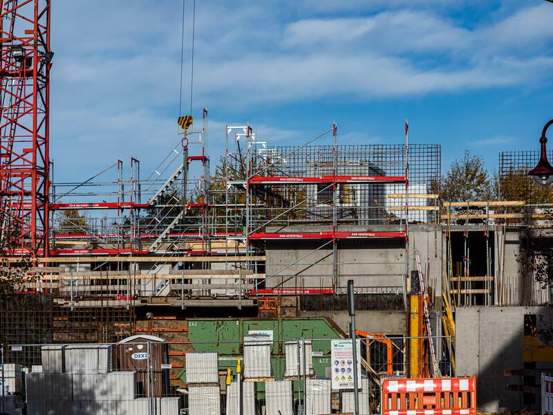 Eine Hausbaustelle, vor der Baumaterial gelagert wird. Ganz links im Bild befindet sich ein roter Mast eines Krans.