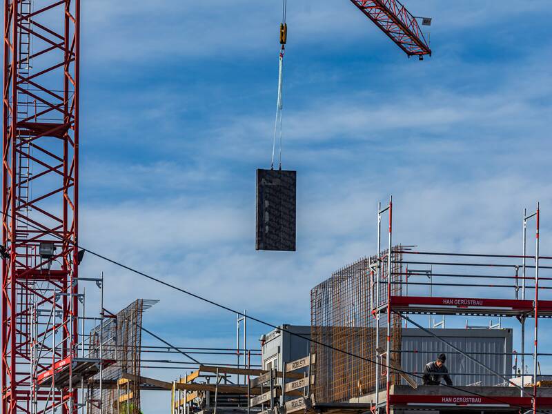 Ein Haus im Rohbau, das eingerüstet ist. Im linken Bildbereich befindet sich ein roten Kran,  welcher gerade Material an die Baustelle befördert.