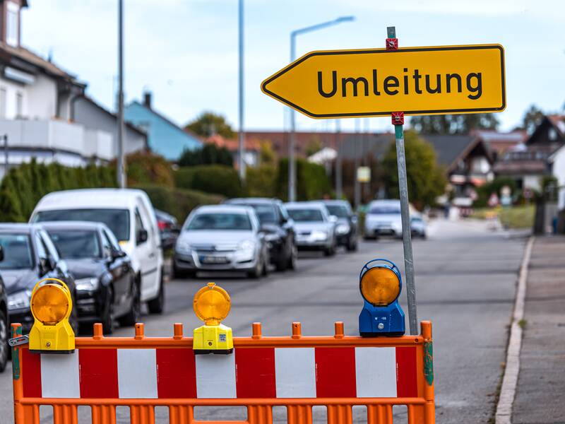 Ein gelbes Umleitungsschild, das in die linke Richtung zeigt. Unter dem Schild steht eine Baustellenabsperrung.