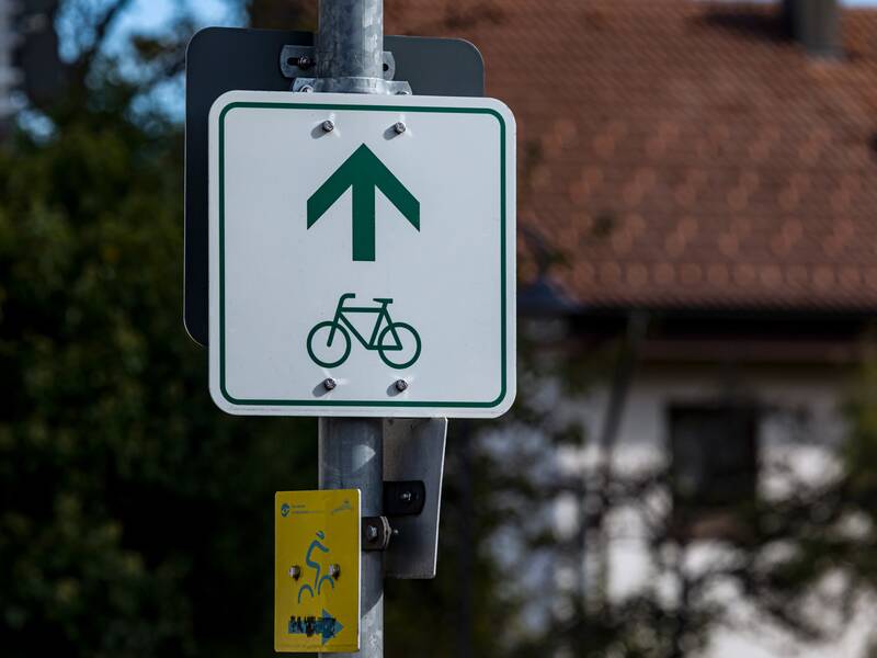 Ein weißes Schild mit einem grünen Fahrrad und Richtungspfeil als Wegweiser für einen Radweg. Das Schild ist an einem Laternenpfosten befestigt.