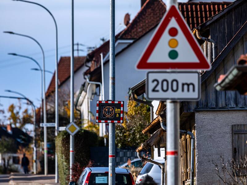 Ein Schild an einem Laternenpfosten mit drei Kreisen in rot, orange und grün, das auf eine Ampel in 200 Metern hinweist. Im Hintergrund sind Häuser und weitere Straßenlaternen.